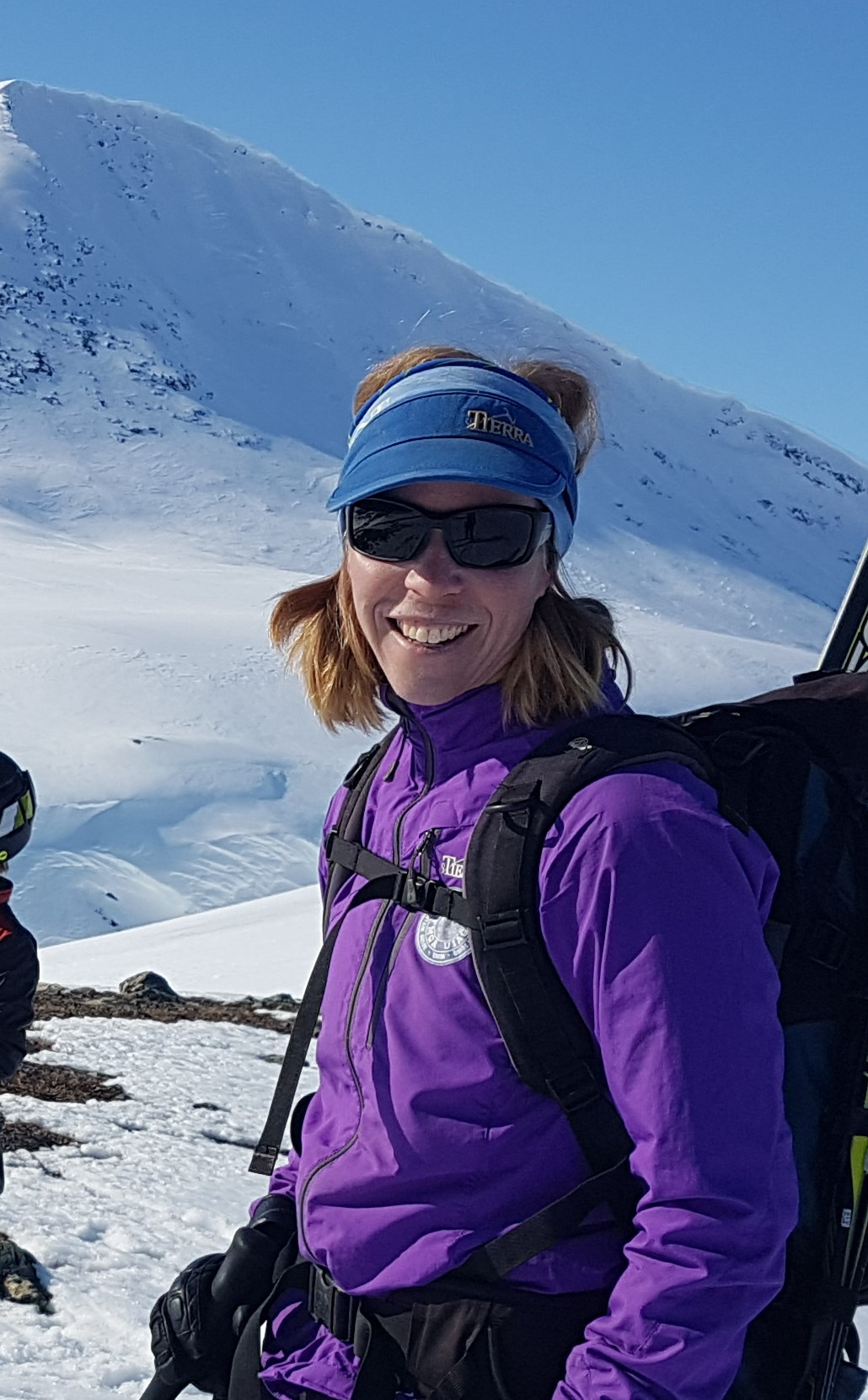 N&auml;rbild p&aring; Sara Widell som st&aring;r framf&ouml;r ett sn&ouml;kl&auml;tt fj&auml;ll ikl&auml;dd en lila jacka, solglas&ouml;gon, svart ryggs&auml;ck och har skidstavar i h&auml;nderna.