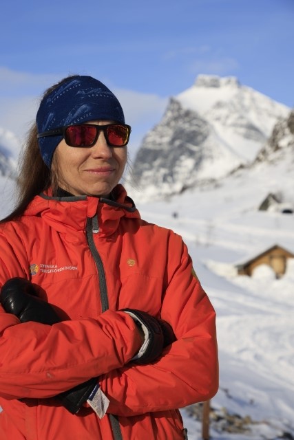 Marit Sarri med långt brunt hår i röd täckjacka och solglasögon. Snöklädda fjäll i bakgrunden.